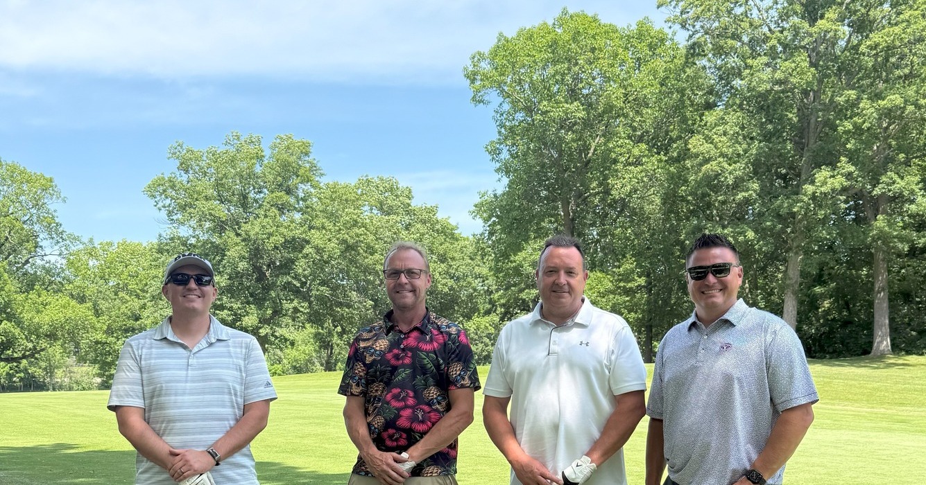 Four men face the camera on a golf course on a sunny day